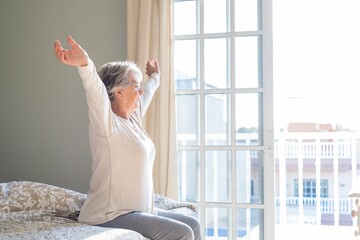 Senior woman sitting on bed with arms wide outstretched and eyes closed in bedroom next to window...