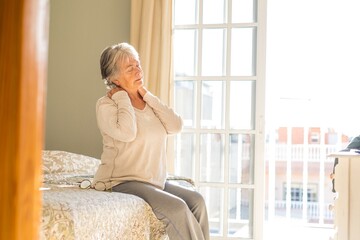 Senior woman suffering from neckache after sleep, rubbing stiff muscles, Old female sitting on bed...