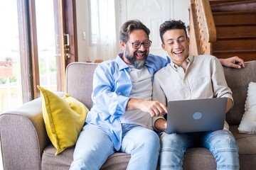 Happy father and son looking at laptop screen while sitting on couch in living room at home. Father with teenage son browsing social media content using laptop in living room - Powered by Adobe