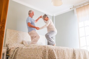 Elderly happy senior couple holding hands and dancing together on bed at home. Carefree active senior heterosexual couple holding hands and dancing together on bed at home - Powered by Adobe