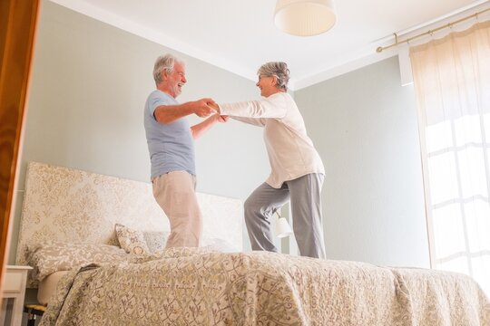 Elderly happy senior couple holding hands and dancing together on bed at home. Carefree active senior heterosexual couple holding hands and dancing together on bed at home - Powered by Adobe