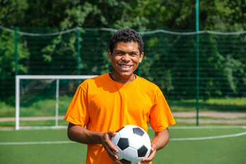 Portrait of young smiling African-American man holding soccer ball while standing on sports court outdoors against soccer goal