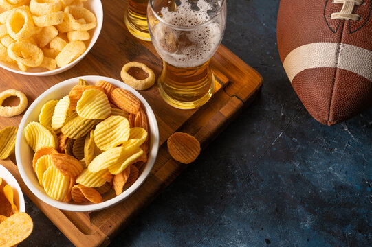 Potato chips, onion rings, lager beer in glasses on a wooden tray, and a baseball glove. Dark blue background. High angle view. Watching sports matches on TV with friends. Rest, relaxation.
