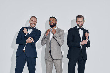 Three handsome young men in suits and bowties looking at camera and smiling while standing against gray background