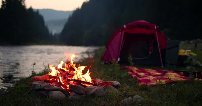 dancing flames of a campfire with tent, river and forest mountain on the background. Travelling and camping concept