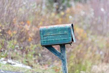 A vintage country style green metal postal mailbox with the door slightly open on a wooden post. The letterbox has colorful autumn trees in the background with orange, green, and yellow leaves.