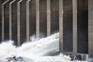 A hydroelectric dam with clear freshwater spilling over the rounded dam edges.  The overspill hits rocks. The water is causing foam to collect at the bottom of the wall and near the large boulders.