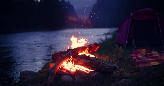dancing flames of a campfire with tent, river and forest mountain on the background. Travelling and camping concept