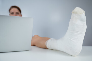 Caucasian woman lifted her leg with plaster to work desk and works on laptop on white background