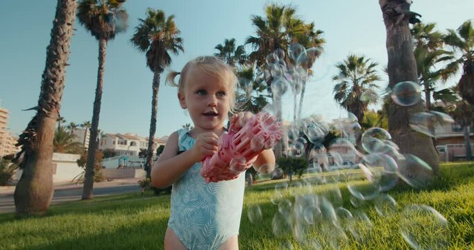 Little and blonde baby girl is playing with bubbles rose color toy gun in public park at summer day on the sunset and smiling with palm trees on background. Shooting to the camera, handheld footage