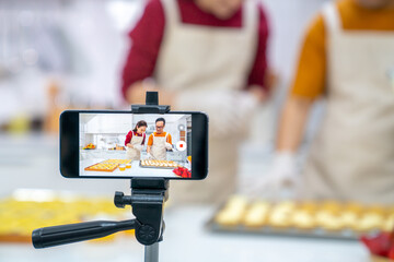 Asian couple bakery shop owner using smartphone with internet vlogging sweet dessert baking on social media together in the kitchen. Small business entrepreneur and online cooking class concept
