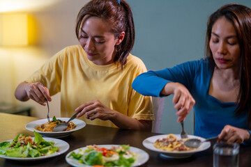 Two Asian woman friends having dinner eating pasta and salad with talking together at home....