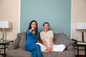 Millennial Asian woman friends sitting on sofa watching movie on tv with eat snack together in living room. Female friendship enjoy and having fun weekend activity lifestyle with home entertainment