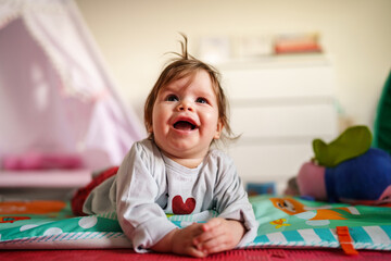 Obraz premium Baby small caucasian girl five months old lying belly down on tummy at home in summer day playing with toy childhood concept looking to the camera alone front view smiling