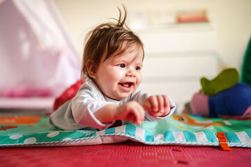 Baby small caucasian girl five months old lying belly down on tummy at home in summer day playing with toy childhood concept looking to the side alone front view smiling