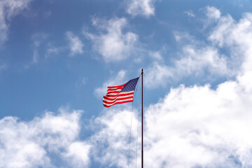 A tattered or weathered American flag on a flagpole with 50 stars and white and red stripes waves and flaps in the wind with torn edges with clouds and blue sky beyond.