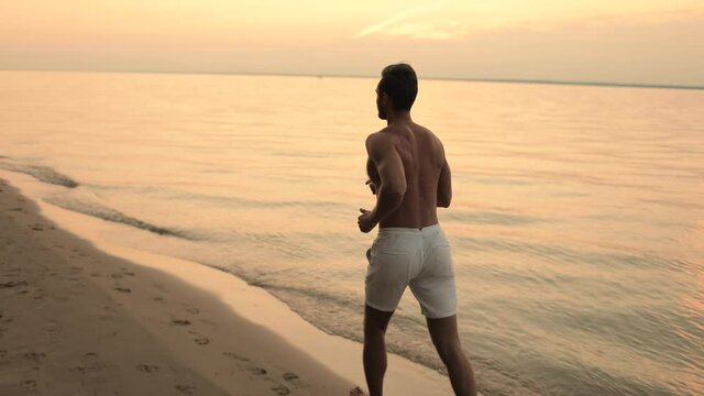 Beautiful gentle sunrise or sunset and a fog. Athletic young man in white shorts running on the sand along the bank. Slow motion. Back view. Healthy lifestyle concept.
