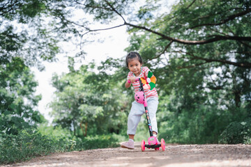 Asia children learn to ride scooters in a park on a summer day. Preschooler girl riding a roller. Kids play outdoors with scooters. Active leisure and outdoor sport for children