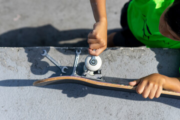 Top view of a concentrated child repairing his skateboard before using it inside the skateboard park on a sunny day. High quality photo