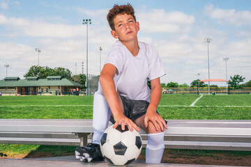 Young male soccer player sitting on bench with ball waiting anxiously to play