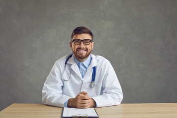 Happy friendly smiling doctor in white coat with stethoscope sitting at desk looking at camera against studio background and looking at camera. Perfect medical healthcare service concept