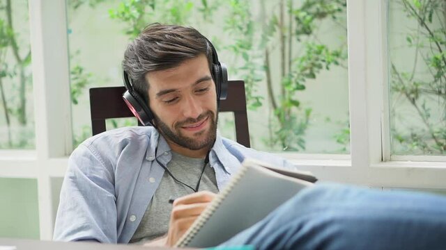 happy young handsome freelancer man resting with feet on desk while sitting on working place writing ideal on book or diary with listen music form headphone in room at home. relaxation work at home .