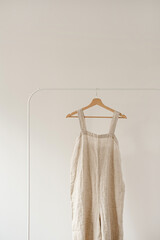 Minimalist wardrobe: Neutral beige colour washed linen overall dress or sundress on hanger against white wall. Aesthetic fashion concept
