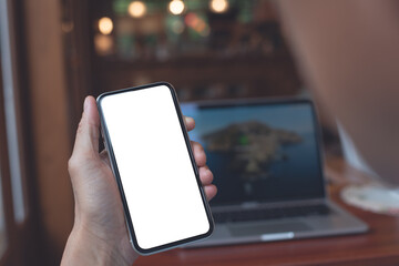 Cell phone mockup image blank screen, man hand holding, using mobile phone with laptop computer on wooden table at coffee shop