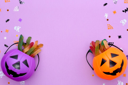 Happy Halloween Concept. Jack O' Lantern Halloween Pumpkin Pails On Purple Background. Orange And Purple Plastic Trick Or Treat Candy Bucket With Gummy Worms Above Frame Made Of Pinata Confetti.