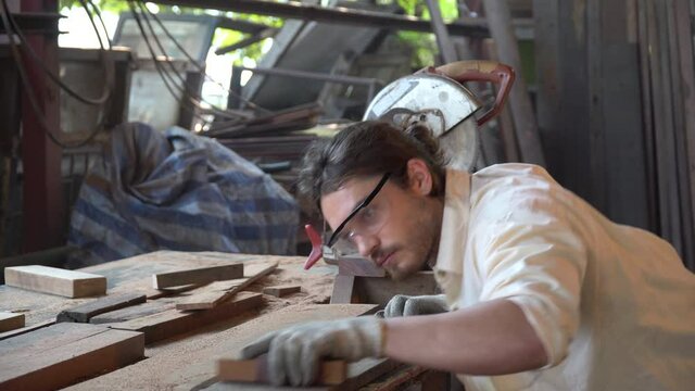 young carpenter man wearing Safety Glasses using sandpaper on a piece of wood and blowing dust in workshop .