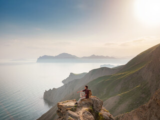 A young Asian woman with a backpack hiking in the summer sits on top of a montage and looks out over a beautiful sea bay landscape. Mountain and coastal travel, freedom and an active lifestyle