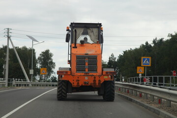 Big heavy 3-axle orange wheeled bulldozer tractor drive on the suburban highway before the pedestrian crossing at summer day, industrial machine traffic on the road