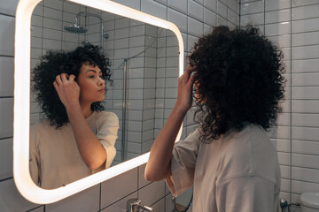 Young pretty woman checking herself in the mirror in modern bathroom. Putting curly hair behind ear.