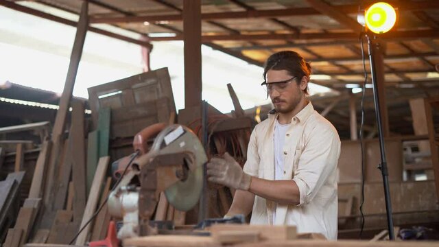 carpenter using circular saw cutting wooden plank in workshop . Young man builder sawing board . craftsman wearing protective safety glasses .