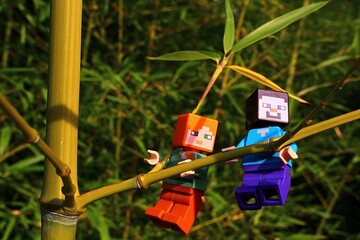 Obraz premium LEGO Minecraft figures of Steve and Alex climbing together on side branch of bamboo plant, genus Phyllostachys, in dense bamboo forest, summer afternon sunshine. 