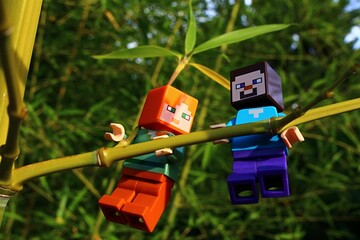 Obraz premium LEGO Minecraft figures of Steve and Alex climbing on side branch of bamboo plant, Phyllostachys genus, sunlit by late afternoon summer sunshine. 