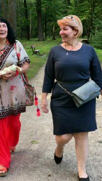 Two mature adult women walking in park talking and smiling. On sunny summer day.