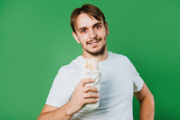 A young man in a white T-shirt on a green background holds lunch, a doner with chicken.