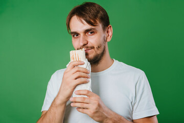 A young man in a white T-shirt on a green background holds lunch, a doner with chicken.