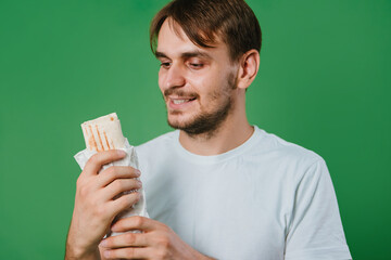A young man in a white T-shirt on a green background holds lunch, a doner with chicken.