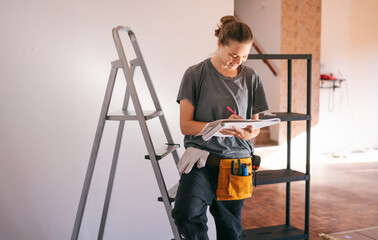 Beautiful young woman professional repairman standing next to a stepladder taking notes while writing down dimensions in a notebook. House renovation concept