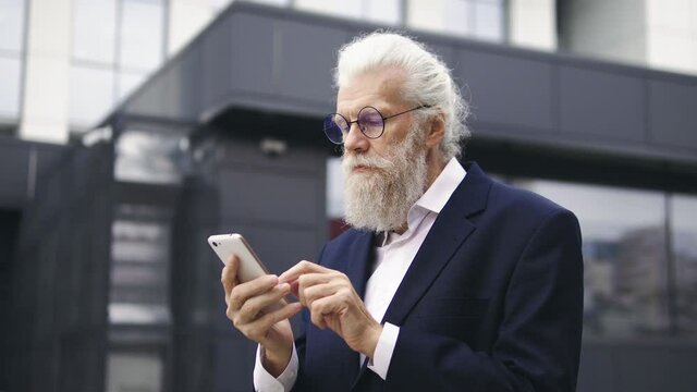 Senior grizzled man scrolling web-pages on smartphone, waiting partners outdoor