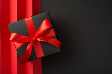 Top view photo of giftbox in black packaging with vivid red ribbon bow and red sheet with vertical...