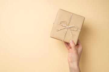 First person top view photo of hand holding craft paper giftbox with twine bow on isolated beige background with empty space