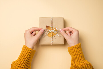 First person top view photo of hands in yellow pullover tying twine bow on craft paper giftbox with yellow autumn leaf on isolated beige background