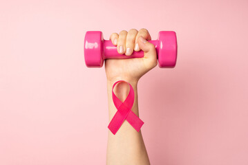 First person top view photo of raised woman's hand holding pink dumbbell and pink ribbon on wrist symbol of breast cancer awareness on isolated pastel pink background