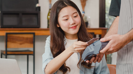 Young Asian freelance women pay contactless credit card at coffee shop. Asian happy men barista waiter wear gray apon holding credit card reader machine for customer can pay with technology in cafe.