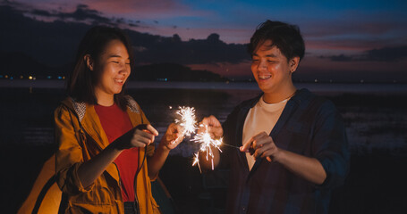 Young asia couple having fun and celebrates with sparklers lights in camping on beach at night. Male and female traveler relax at campsite. Outdoor activity, adventure travel, or holiday vacation.