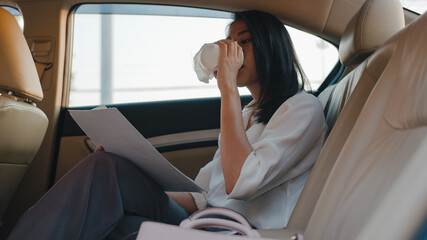 Successful young Asia businesswoman in fashion office clothes working and drinking disposable paper cup of hot drink in back seat of car in urban modern city in morning. Business on the go concept.