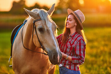 Young cute happy joyful satisfied smiling cowgirl with beautiful blond palomino horse at meadow at...